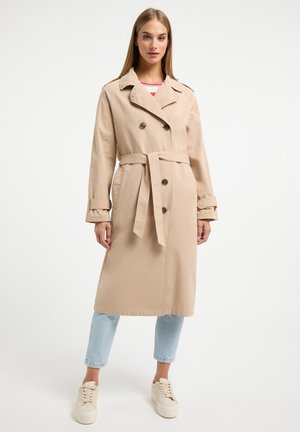 Beige trenchcoat met dubbele knopen, ceintuur in de taille en opgerolde mouwen, gecombineerd met lichtblauwe cropped jeans en crèmekleurige sneakers.