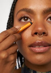 Orange eyelinerpenna applicerad på det undre ögonlocket, hålls av en hand. Modellen har flätat hår, en gyllene septumring och glansiga läppar.