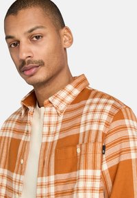 Camisa de franela de cuadros naranja y crema con cuello abotonado, que presenta un bolsillo en el pecho y botones blancos. Textura suave.