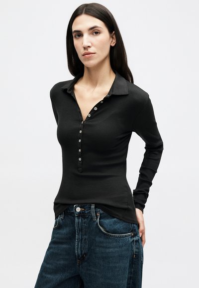 Femme aux longs cheveux foncés portant un haut noir à manches longues boutonné et un jean bleu, debout devant un fond blanc uni.