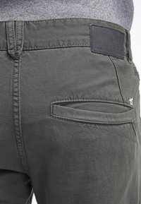 Zadní kapsa tmavě šedých kalhot s poutkem na opasek, viditelným prošitím a černou značkovou štítkem na pásku, nošena se šedou košilí.