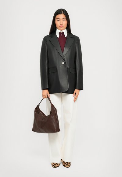 Woman standing wearing a black leather blazer, burgundy zip-up top, white collar, white pants, leopard-print shoes, holding a brown leather handbag.