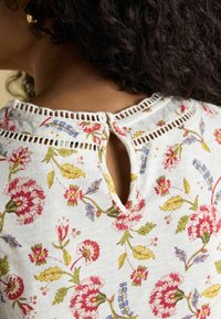 Biała bluzka w kwiatowy wzór z kolorowymi kwiatami, motywami liści i dekoltem w kształcie łezki z guzikem. Wykonana z miękkiej, fakturowanej tkaniny.