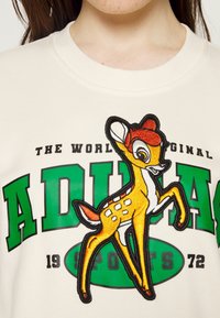 Krämfärgad sweatshirt med en livfull broderad hjort i orange och gult, tillsammans med fet grön och svart text som säger "VÄRLDENS ORIGINAL ADIDAS SPORTS 1972."