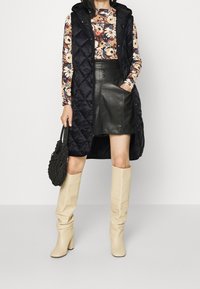 Černá prošívaná vesta s kapucí přes květinové tričko s dlouhým rukávem, spárovaná s černou koženou mini sukní a béžovými kozačkami po kolena. Černá kabelka.