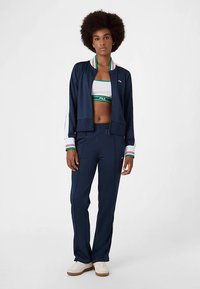 Laivastonsininen vetoketjutakki, jossa on vihreä ja punainen raidallinen kaulus, yhdistetty sopivaan istuvaan lyhytpaitaiseen toppiin ja väljien housujen kanssa, käytetty valkoisten tennarien kanssa.