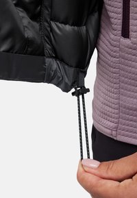 Schwarze Steppjacke mit einem gesteppten Innenfutter, das eine verstellbare Kordel zeigt, die in einer Hand gehalten wird. Eine lila Unterwäsche ist darunter sichtbar.