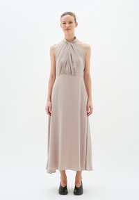 Maxikleid mit Halterhals aus weichem beigen Stoff, mit plissiertem Oberteil, fließendem Rock und schwarzen spitzen Ballerinas. Sanfte Textur, schlichtes Design.