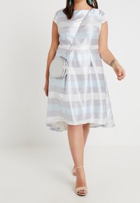 Ljusblå och grå randig klänning med en åtsittande bodice och en utsvängd kjol. Har korta ärmar och en glänsande yta. Kombineras med en rund silverväska.