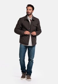 Gequilted donkerbruine jas met een kraag, voorkant zak en drukknoopsluiting, gedragen over een gestreept shirt, gecombineerd met jeans en casual schoenen.