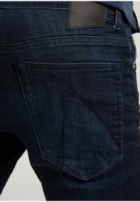 Tmavě modré džínové kalhoty s texturovanou tkaninou, výrazným designem zadních kapes a koženou značkovou nášivkou na pasu.