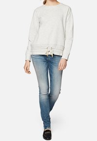 Sudadera gris claro con cuello redondo y dobladillo con cordón, combinada con jeans azul medio y zapatillas negras sin cordones.