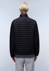 Personne blonde portant une veste matelassée noire, un pull gris et un pantalon gris clair, vue de dos sur un fond uni.