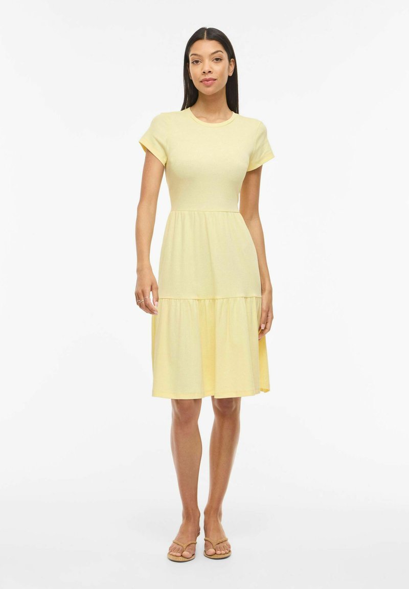 Sieviete stāv, valkājot īss piedurkņu, gaiši dzeltenu ceļgalu garuma kleitu ar kārtainu svārku un plakanām sandales uz baltas fona.