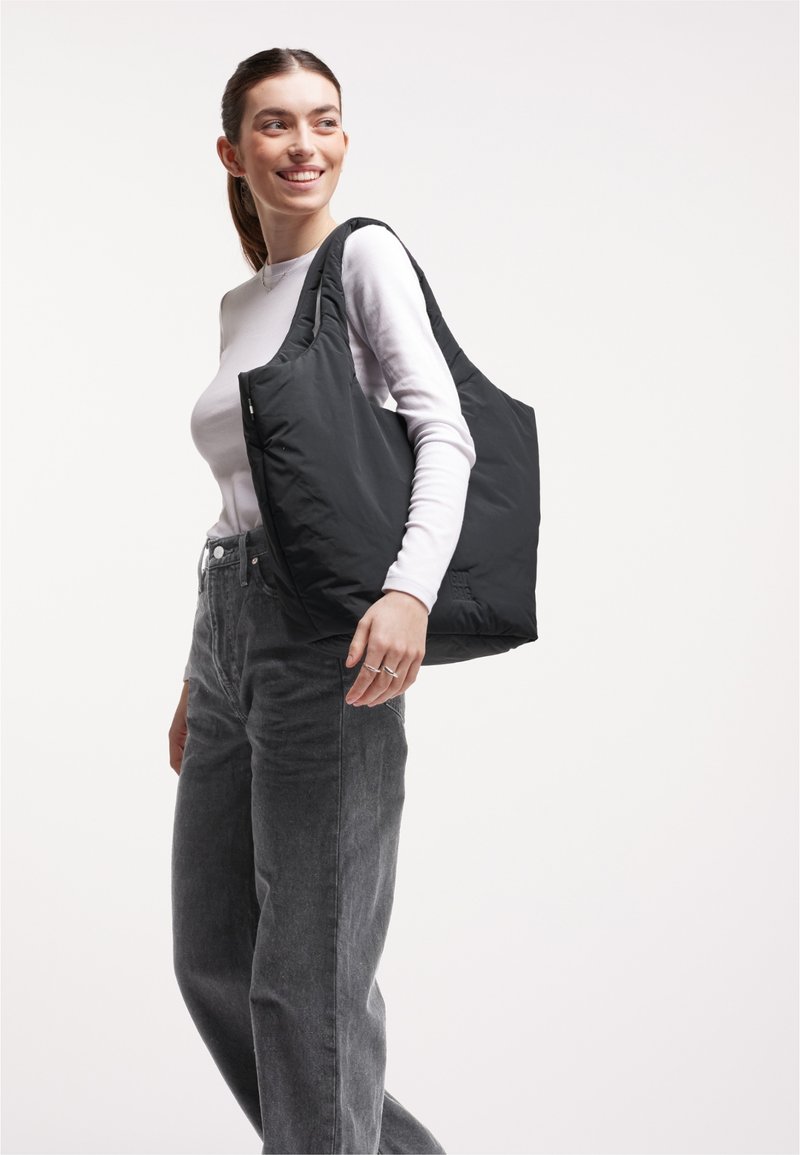 Femme souriante, portant une chemise blanche à manches longues et un jean gris, portant un grand sac fourre-tout noir rembourré sur l'épaule, sur un fond uni.