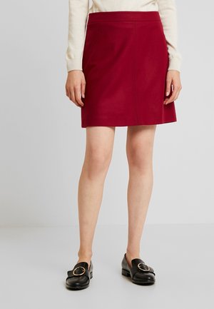 Femme portant une jupe rouge mi-genoux, un haut crème à manches longues et des mocassins noirs avec de grandes boucles dorées, debout sur un fond uni.