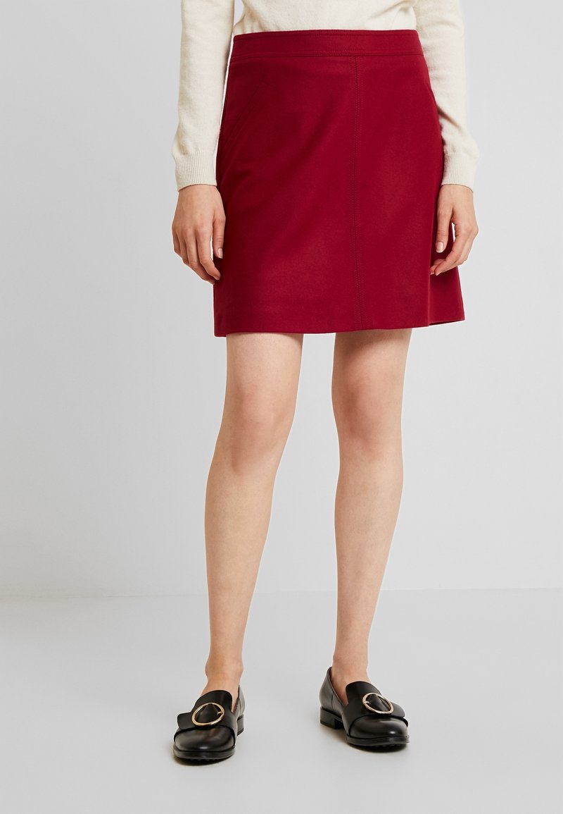 Femme portant une jupe rouge mi-genoux, un haut crème à manches longues et des mocassins noirs avec de grandes boucles dorées, debout sur un fond uni.