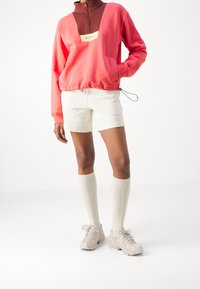 Felpa zip-up corallo e marrone con un grande taglio, abbinata a pantaloni corti bianchi e calze al ginocchio a coste. Le scarpe sono di stile atletico beige.