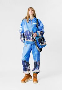 Sudadera gráfica azul con cuello de media cremallera, pantalones a juego y botas marrones bicolor. Presenta un estampado de paisaje y detalles en contraste.