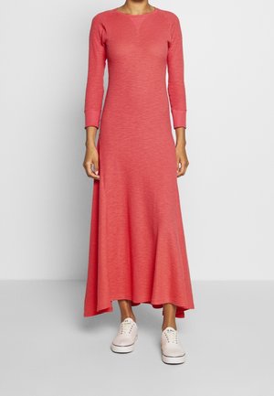 Kobieta ubrana w koralową sukienkę z długim rękawem sięgającą kostek z rozszerzanym dołem oraz białe tenisówki z sznurowadłami na jednolitym jasnoszarym tle.