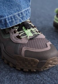 Sports shoe with a textured brown upper, green laces, and a toggle closure. Features a thick sole with a checkerboard pattern.