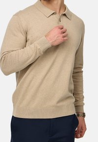 Suéter de polo de punto beige con cuello, dos botones y puños acanalados; textura suave. Llevado con pantalones azul marino.