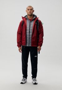 Jachetă impermeabilă roșie peste un vestă puf gri, pantaloni negri cu logo, adidași albi. Modelul poartă un rucsac cu accente verzi.