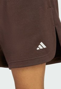 Hnědé sportovní šortky s malým bílým logem adidas poblíž lemu, nošené na stehně osoby na jednolitém pozadí.