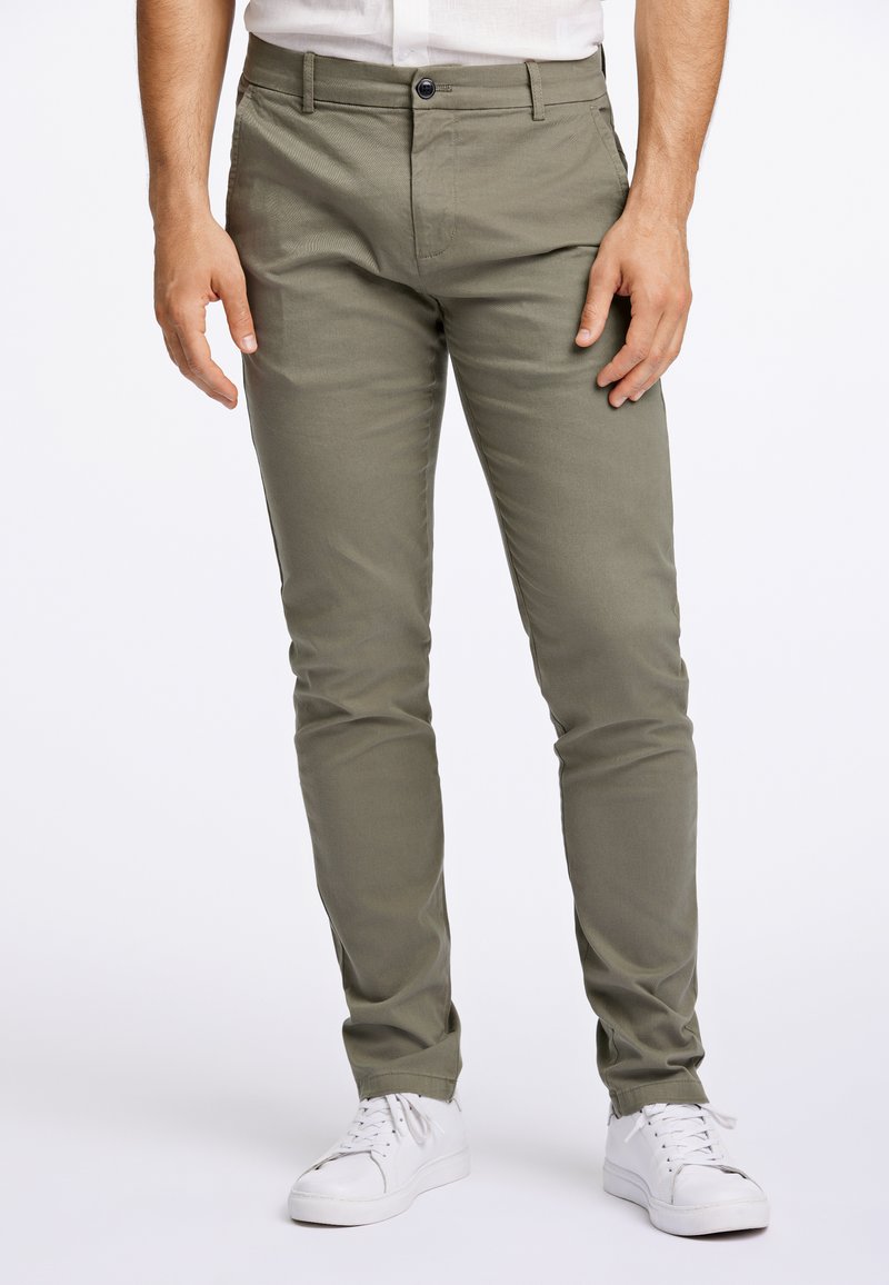 Slim-Fit, olivgrüne Chinos aus Baumwollmischgewebe, mit Knopfverschluss, Seitentaschen und passender Steppnaht zu weißen Sneakers.