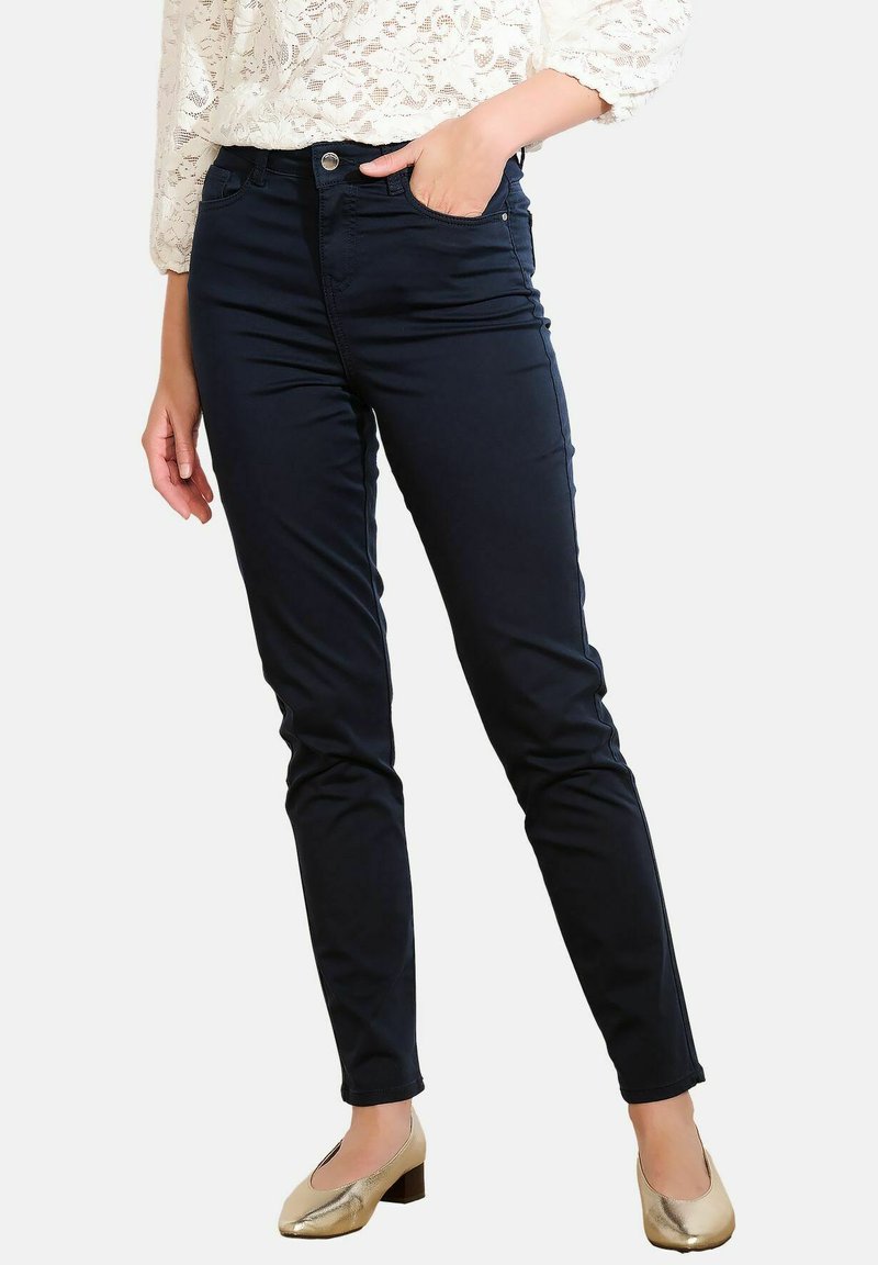 Žena v černých slim-fit kalhotách, bílé krajkové blůze a zlatých nízkých podpatcích, stojící s jednou rukou v kapse.