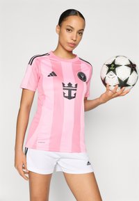 Rózsaszín csíkos focimez fekete díszítéssel és logóval, fehér nadrággal. A játékos egy mintás focilabdát tart.