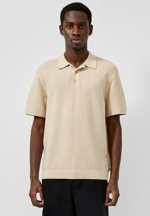 Młody mężczyzna o ciemnej karnacji, ubrany w beżową dzianinową koszulkę polo z krótkim rękawem i czarne spodnie, stoi na tle jednolitego jasnego tła.