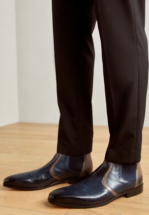 Stivali Chelsea in pelle con una texture in coccodrillo blu scuro, accenti marroni a contrasto e un design snodato, abbinati a pantaloni neri su misura.