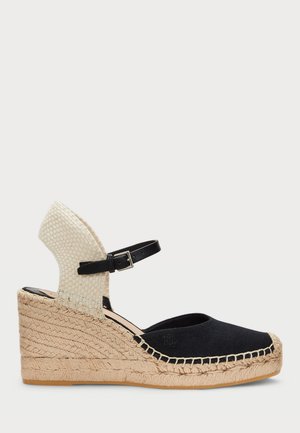 Black canvas espadrille with a woven beige ankle strap, jute-wrapped wedge heel, and rubber sole. Features a rounded toe and contrasting stitching.