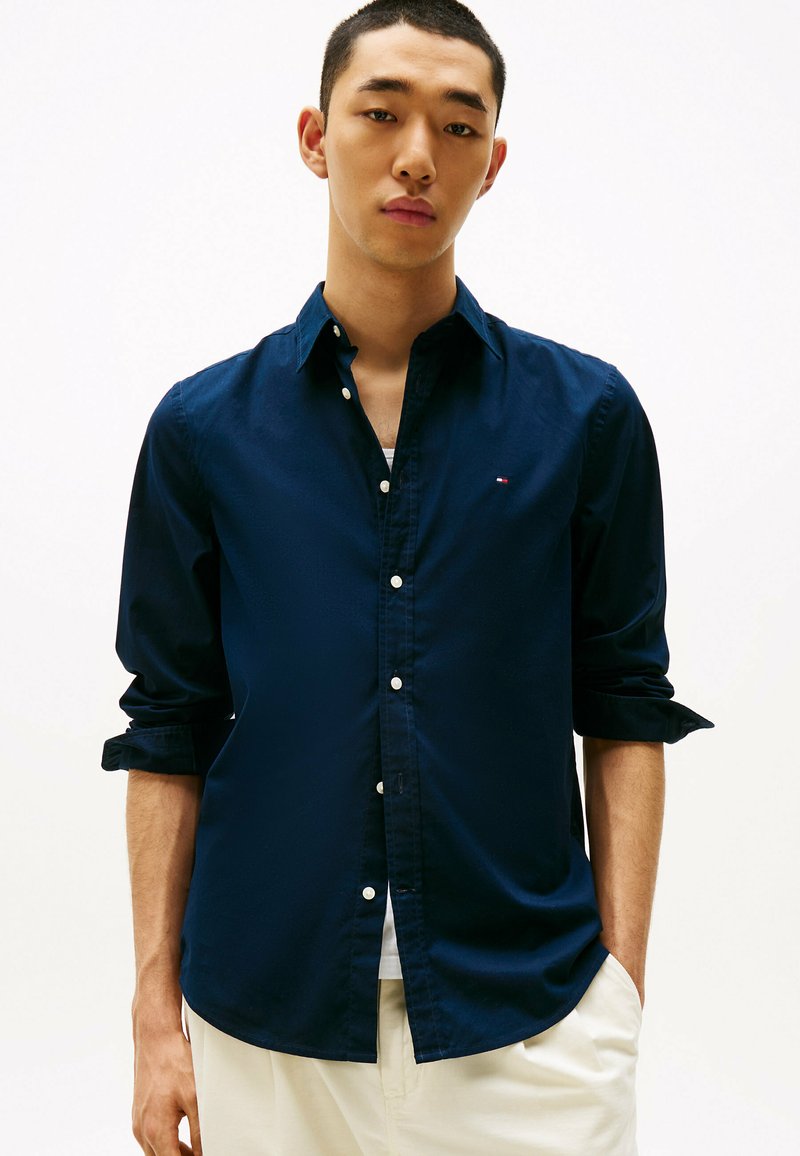 Camisa de botonadura en azul marino, hecha de tejido trenzado, con un cuello clásico, mangas largas con puños enrollados y un pequeño detalle de logo.