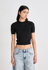 Svart ribbet kortermet topp med rund halslinning og en kort design. Har en liten rød og hvit logo på brystet. Denimsjeans med en falmet look.