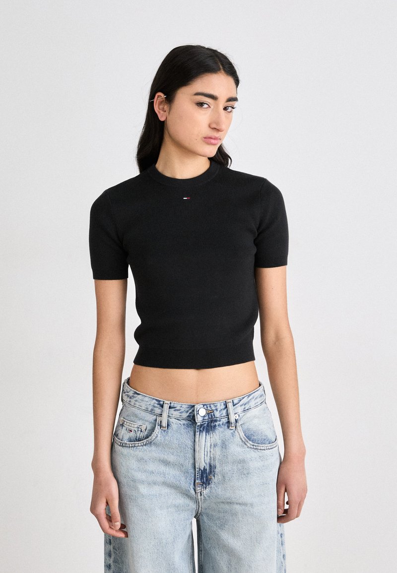 Svart ribbet kortermet topp med rund halslinning og en kort design. Har en liten rød og hvit logo på brystet. Denimsjeans med en falmet look.