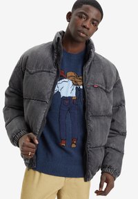 Puffjakke i mørkegrå denim med glidelås foran og elastiske mansjetter. Underneath har en blå genser et fargerikt strikket mønster.