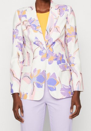 Woman wearing a white blazer with purple and yellow floral patterns, a yellow top, and light purple pants, standing against a plain background.