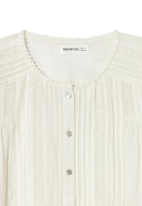 Weiße Bluse mit rundem Ausschnitt, zartem Spitzenbesatz, vertikalen Bundfalten und floralen Stickereidetails sowie kleinen Perlenknöpfen vorne.