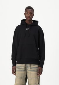Hoodie noir en coton, avec une poche frontale, des cordons de serrage et un petit logo sur la poitrine. Associé à un pantalon beige à motifs.