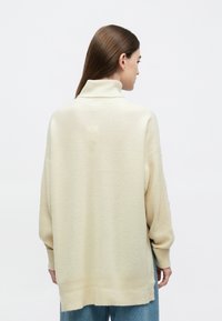 Kreminės spalvos didelio dydžio megztinis su aukštu apykakle, riboto rašto tekstūra ir nuleistais pečiais, derinamas su languotais džinsais laisvo silueto.