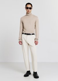 Beige Rollkragenpullover, cremefarbene maßgeschneiderte Hose und schwarze Knöchel-Stiefel. Das Model trägt schwarze rechteckige Sonnenbrillen mit einem minimalistischen Design.