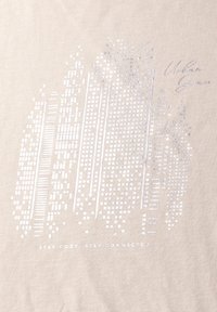 Camiseta beige con un diseño gráfico plateado que presenta líneas verticales y puntos, con el texto "MANTENTE CÓMODO, MANTENTE CONECTADO" y una firma manuscrita.