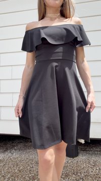 Kvinne iført en svart off-shoulder kjole med volangdetalj, stående utendørs på grus foran en hvit kledningvegg.