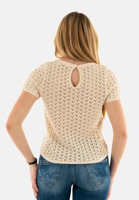 Top corto de punto beige con un patrón de encaje, que presenta una abertura en la parte posterior y bordes acanalados. Combinado con unos jeans ajustados azules.