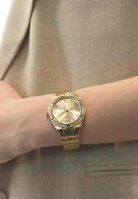 Reloj de pulsera dorado con caja redonda, esfera dorada texturizada, números romanos y acentos decorativos. La pulsera de metal añade elegancia.