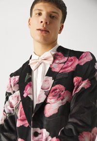 Black blazer with large pink rose print, featuring a satin pink bow tie and a white shirt. The fabric has a smooth, sleek texture.