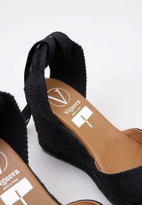 Sandalias de cuña de tejido negro trenzado con una plantilla color tostado, que presentan un logo y una tira al tobillo. El tacón tiene textura para ofrecer mejor agarre.