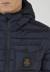 Giacca puffy blu navy con cappuccio, caratterizzata da una zip frontale, tasca sul petto e una toppa in gomma con logo. Design a trama trapuntata.